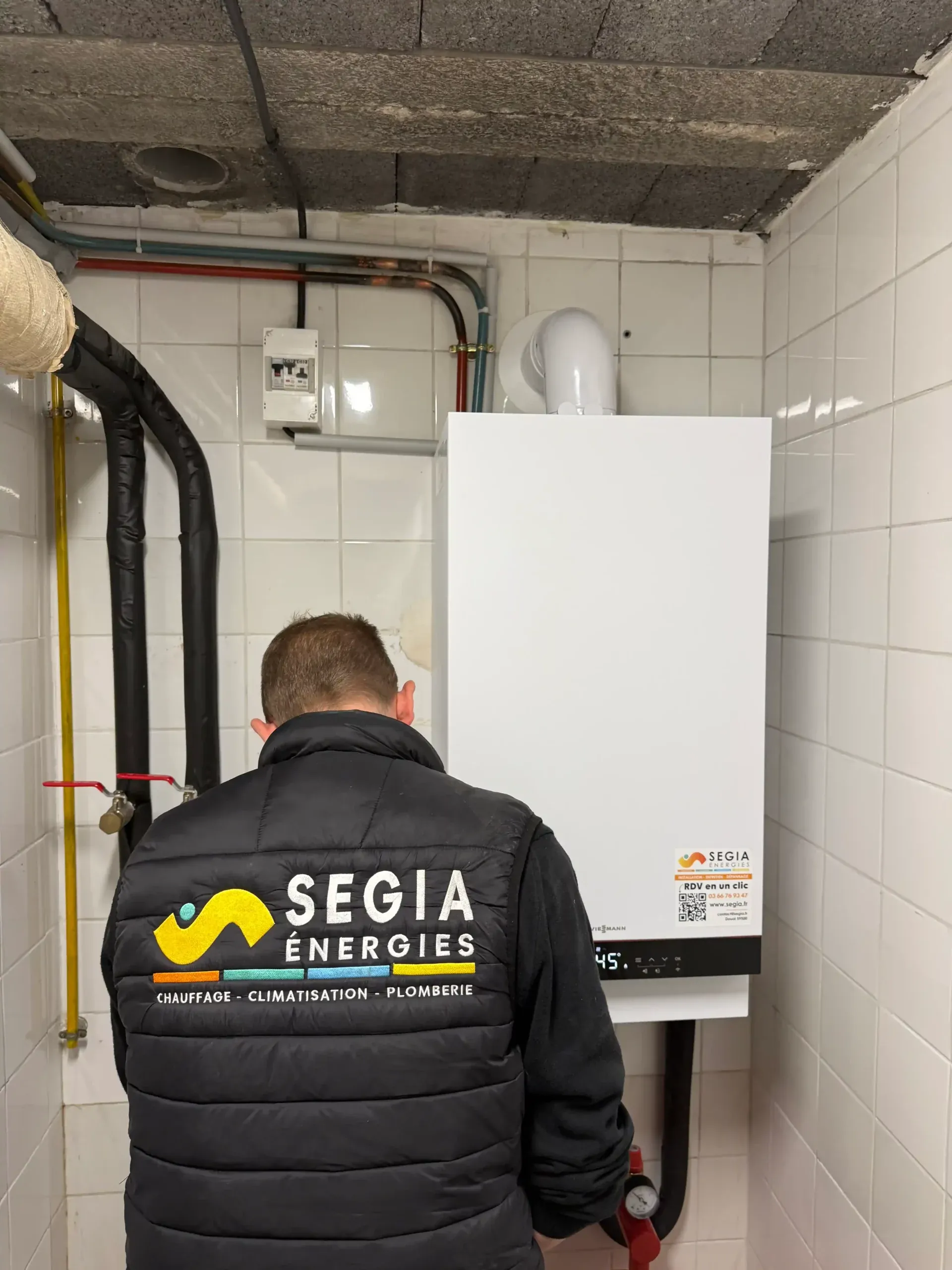 Technicien SEGIA de dos devant une chaudière murale Viessmann blanche posée dans un local carrelé, avec ses tuyaux de gaz et de chauffage